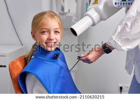 Young child girl next to dental 3D x-ray digital scanner panorama machine in clinic. Portrait of caucasian cute girl looking at camera, cropped dentist is preparing equipment to take x-ray picture
