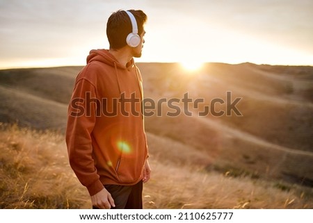 Side View On Handsome Caucasian Guy In Headphones Looking Back At Sunrise, Having Rest After Jogging, Running, Sport Outdoors. Fit Guy In Casual Wear Hoodie During Training, Alone Royalty-Free Stock Photo #2110625777