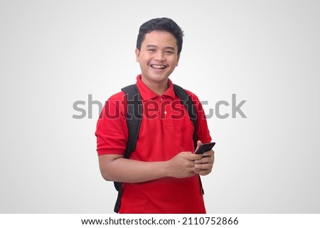 Portrait of excited Asian student man in red polo shirt wearing a backpack, looking at camera and holding mobile phone. Isolated image on white background