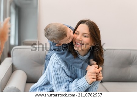 A young mother and her son sit on a sofa at home, hug and kiss. Kiss on the cheek.