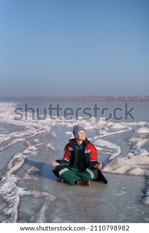 A woman in a colorful thermal costume enjoying winter while sitting in a meditation pose on a frozen lake with blue sky in the background. Freedom, peace, quiet, serenity