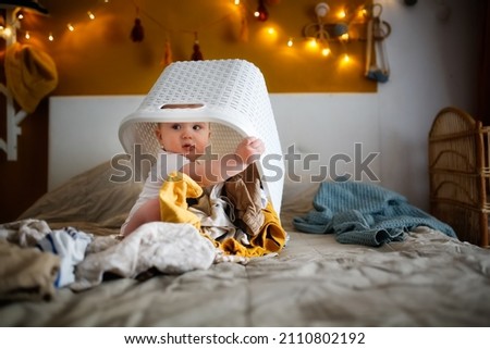 funny european chubby kid playing with laundry basket on bed in cozy bedroom, mom help and home routines, motherhood and baby care Royalty-Free Stock Photo #2110802192