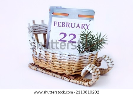 Calendar for February 22: a decorative sleigh with a loose-leaf calendar inside, the name of the month in English, the number 22, a fir branch, a white background