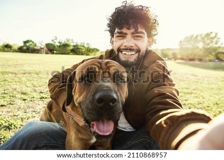Young happy man taking selfie with his dog in a park - Smiling guy and puppy having fun together outdoor - Friendship and love between humans and animals concept 