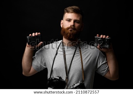 Professional photographer in a grey t-shirt with a bunch of different cameras in a hands and on a shoulder looking on a camera and ready for make a good shoot on a black background.