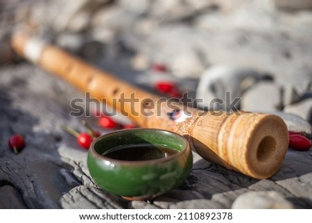 The flute of the Shakuhachi lies diagonally. Near in the foreground is a green mug. It is filled with Chinese Pu-erh tea. The composition lies on gray stones and next to it are wild rose berries
