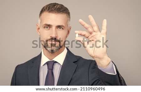 mature business man with grizzled hair in suit hold pill in hand with ok gesture, energy remedy