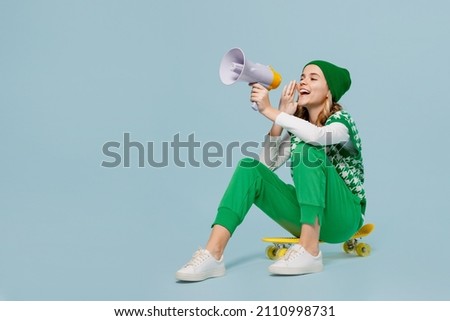 Full size body length excited young brunette girl teen student wears checkered green vest hat sit on skateboard hold scream in megaphone isolated on plain pastel light blue background studio portrait Royalty-Free Stock Photo #2110998731