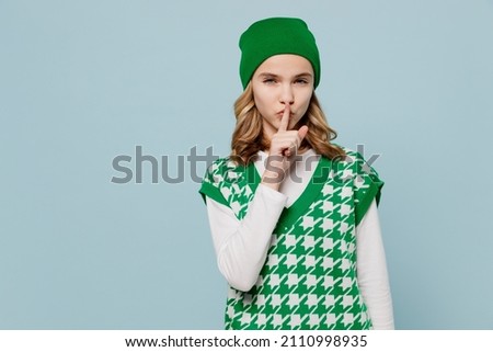 Secret fascinating fun young brunette girl teen student wears checkered green vest hat say hush be quiet with finger on lips shhh gesture isolated on plain pastel light blue background studio portrait