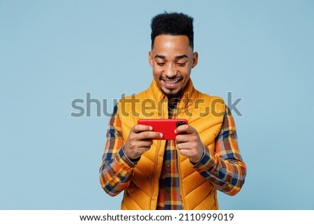 Fun young black man 20s wears yellow waistcoat shirt using play racing app on mobile cell phone hold gadget smartphone for pc video games isolated on plain pastel light blue background studio portrait