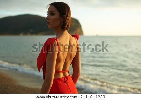 Awesome young woman in red fashionable clothes on the sand beach. Beautiful sea landscape on background. Brunette with big eyes and lips. Tan and slim. Summer, sunset. Colorful and warm picture.