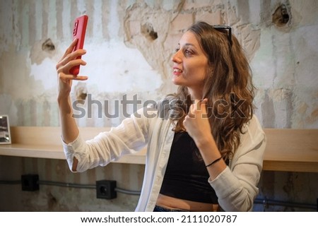 Female portrait of young active girl in white shirt taking a photo of herself indoors on loft wall background, selfie time, happy people concept.