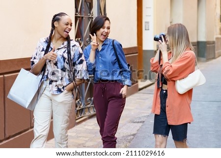 Joyful young multiracial female tourists, smiling and winking while showing v sign and thumb up gesture, during walk on city street with female friend taking picture on photo camera
