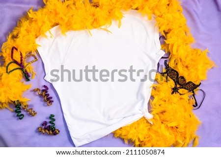 Mockup white t-shirt decorated yellow feather boa and carnival mask. Mardy Gras apparel flatlay on fabric lilac background, flat lay, top view, copy space.