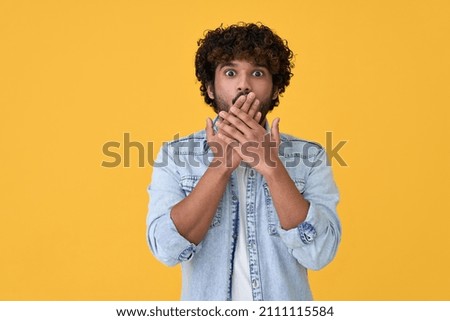 Shocked surprised young indian man looking at camera with omg face expression covering mouth with hands feeling amazed speachless silent reaction standing isolated on yellow background.