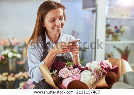 Portrait of female florist taking picture of freshly made bouquet on smart phone, creating content for social media. Small local business, self employed, blogger concept