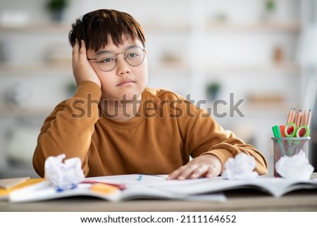 Cute overweight japanese boy teenager doing school project at home, sitting at desk with books and notebooks, got tired and bored, looking for creative solutions, closeup photo, copy space Royalty-Free Stock Photo #2111164652