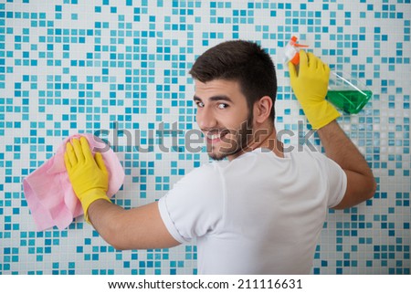 Half-length portrait of young smiling dark-haired janitor wearing white shirt and yellow rubber gloves standing back in the bathroom and cleaning it with the rag and detersive