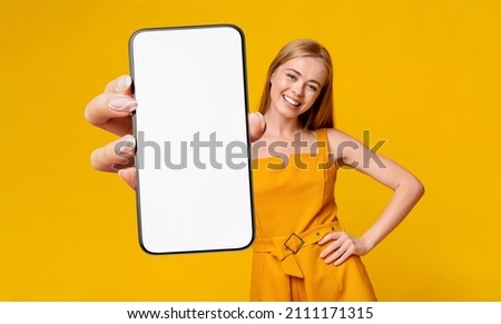 Recommendation. Portrait of excited young lady holding big smartphone with white blank screen in hand, showing device close up to camera. Gadget with empty free space for mock up, yellow orange wall
