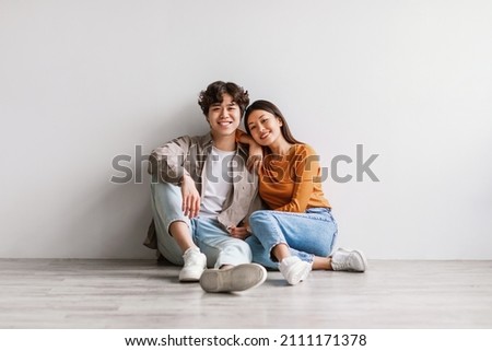 Full length portrait of cheerful young Asian couple in casual outfits sitting on floor and looking at camera against white studio wall. Millennial spouses posing and smiling together