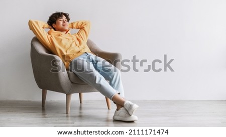 Peaceful young Asian man relaxing in cozy armchair, being lazy, having break against white studio wall, panorama with free space. Handsome millennial guy resting with hands behind head Royalty-Free Stock Photo #2111171474