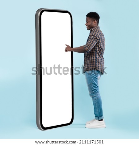Full Body Length Back View Of Black Man Using Big Smartphone With Blank White Screen Touching Huge Display Panel With Finger, Cheerful Guy Standing On Blue Background, Ordering Food Delivery, Mock Up Royalty-Free Stock Photo #2111171501