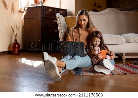 Modern Lifestyle. Portrait of young lady and her little daughter holding and using different electronic gadgets while sitting on the floor in living room, mom working on pc, girl playing with cell