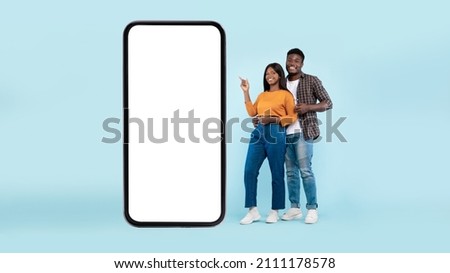Great Offer. Excited couple hugging pointing at big giant smartphone with mock up copy space, promoting app or website, advertising product or service, blue studio wall, banner, full body length