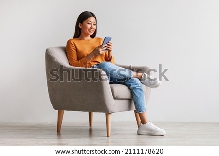 Charming young Asian woman using cellphone, communicating on web, working or learning online, sitting in armchair against white wall, free space. Lovely millennial lady chatting on smartphone Royalty-Free Stock Photo #2111178620
