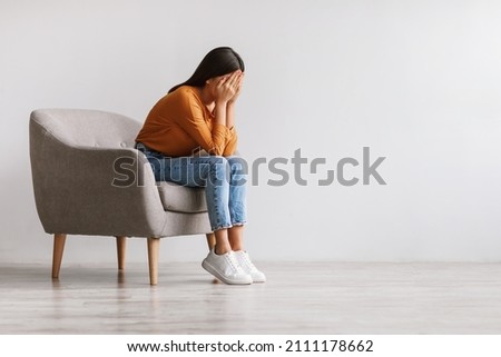 Young Asian woman burying her face in hands and crying, feeling depressed, sitting in armchair against white studio wall, copy space. Millennial lady having mental problems or mood disorder Royalty-Free Stock Photo #2111178662