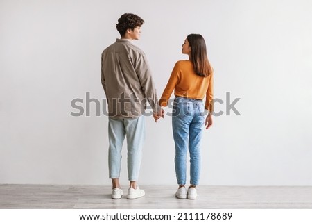Full length of young Asian man and woman standing with their backs to camera, looking at each other and holding hands against white studio wall. Millennial couple expressing affection Royalty-Free Stock Photo #2111178689