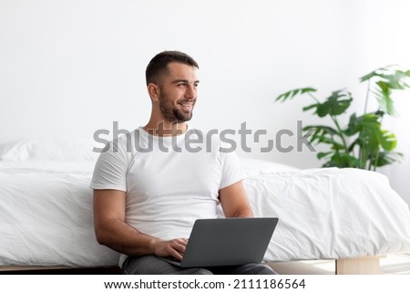 Cheerful young handsome guy with beard work on laptop and look at empty space in minimalist bedroom interior, free space. Idea creation, success business and freelance at home due covid-19 pandemic
