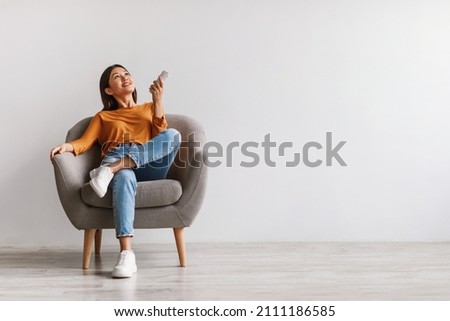 Split system. Happy young Asian woman with remote relaxing under air conditioner, sitting in armchair against white studio wall, mockup. Happy millennial lady using climate control Royalty-Free Stock Photo #2111186585