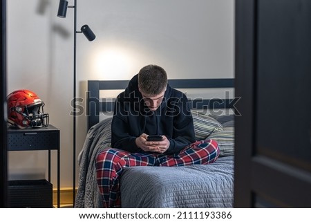 A teenager guy sits in a room on a bed and uses a smartphone.