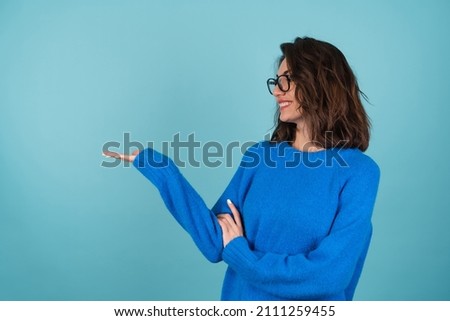 A woman in a blue knitted sweater, curly short hair, glasses on her eyes, smiles cheerfully, points to an empty space, holds in her palm