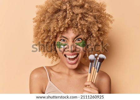 Outraged curly haired young woman screams angrily applies green collagen patches under eyes holds cosmetic brushes going to apply makeup isolated over beige background. Beauty procedures concept Royalty-Free Stock Photo #2111260925