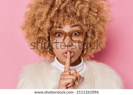Surprised woman with curly bushy hair makes silence gesture tells secret wears spectacles and white fur coat isolated over pink background. Hush do not spread rumors please. Body language concept