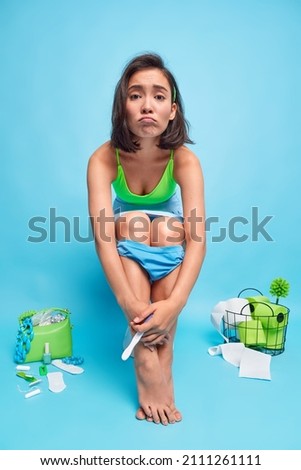 Unhappy young Asian woman poses on toilet bowl with pregnancy test waits for result has panties pulled down on legs surrounded by hygiene items isolated over blue background. Fertility concept