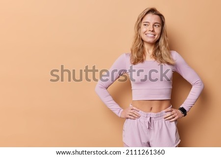Young fit fair haired woman being in good physical shape keeps hands on waist dressed in sportswear smiles happily concentrated away ready for training isolated over beige background blank copy space Royalty-Free Stock Photo #2111261360
