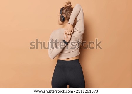 Photo of slim flexible woman clutches hands behind back has hair combed in bun dressed in sportswear does exercises in gym listens music via stereo headphones isolated over beige background. Royalty-Free Stock Photo #2111261393