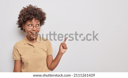 Attractive smiling young curly woman points index fnger at upper right corner demonstrates promotio gives way somewhere wears round transparent glasses and t shirt isolated over white background.