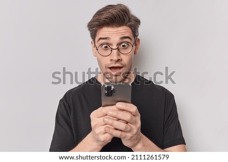 Stupefied European man stares at smartphone screen reads strange message found out shocking truth reacts to unexpected news wears round spectacles isolated over white background. Omg concept Royalty-Free Stock Photo #2111261579