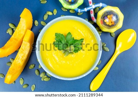 Pumpkin soup puree for the baby in a white bowl, pumpkin slices and seeds, a spoon and a rattle toy on a blue background. Close-up. Baby nutrition, the first complementary food. Royalty-Free Stock Photo #2111289344
