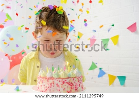 The child blows out the candles on the birthday cake. Boy 5 years old in a festive setting. Birthday, confetti fall on the boy. Horizontal photo. Royalty-Free Stock Photo #2111295749