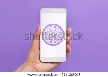 Woman's hand holding mobile phone with astrology application and zodiac circle on white screen isolated on lilac background