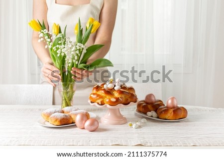 Happy Easter! Woman preparing table with Easter pastry, eggs, candies and spring flowers for holiday. Light background. Copy space. Royalty-Free Stock Photo #2111375774