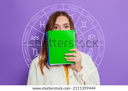 Shocked young woman holding white poster in her hands with zodiac astrological circle isolated on lilac background