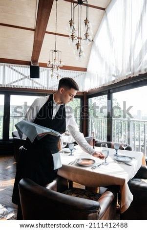A young male waiter in a stylish uniform is engaged in serving the table in a beautiful gourmet restaurant. A high-level restaurant. Table service in the restaurant. Royalty-Free Stock Photo #2111412488