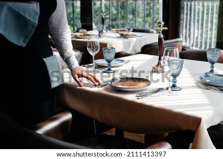 A young waiter in a stylish uniform is engaged in serving the table in a beautiful gourmet restaurant close-up. Table service in the restaurant. Royalty-Free Stock Photo #2111413397