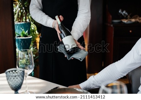A young sommelier in a stylish uniform demonstrates and offers the client fine wine in the restaurant. Customer service. Table setting in a fine restaurant. Close-up. Royalty-Free Stock Photo #2111413412